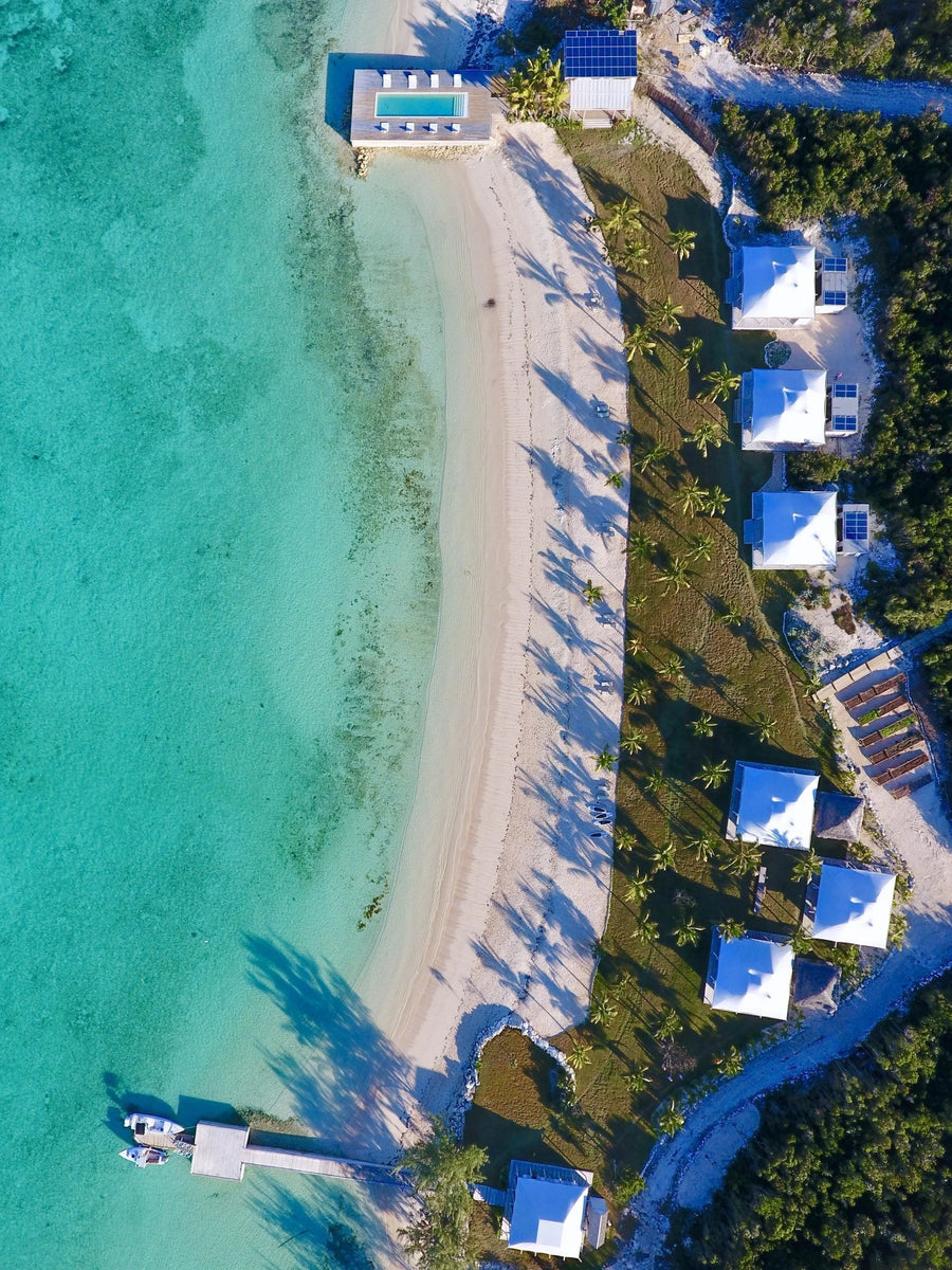 Aerial view of a tropical beachfront resort with turquoise water, a curved white sand beach, palm trees, and several white-roofed villas along the shoreline.