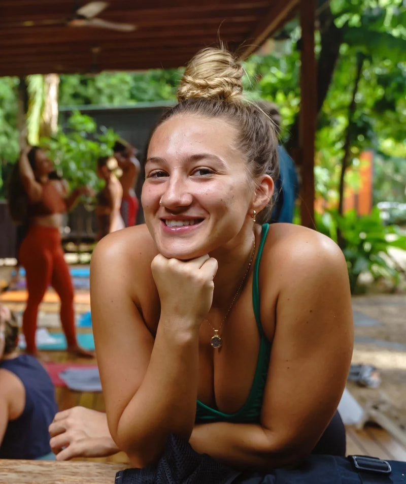 A smiling woman with her hair in a bun leans forward in a tropical outdoor setting, wearing a dark green tank top and a necklace. Other people and yoga mats are visible in the background under a wooden canopy.